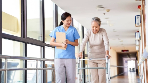 Bir bakıcı, yürüteçli yaşlı bir kadına eşlik ediyor.