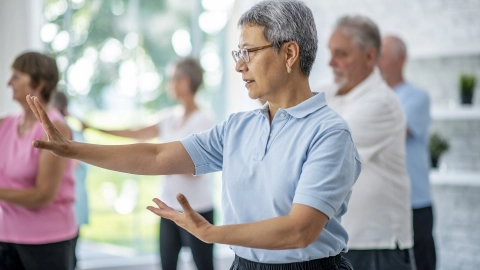 Иммунитет: несколько человек стоят в светлой комнате и выполняют физические упражнения. На переднем плане пожилая женщина, выполняющая движения обеими руками.