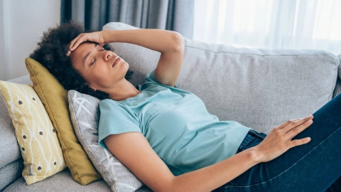 Woman lying on a couch supported on a few pillows. She is clutching her head and looks exhausted.