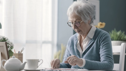 Пожилая женщина сидит за столом в светлой, уютной комнате и собирает пазл. Перед ней на столе стоят цветы, рамка для картины, чайник и чашка.