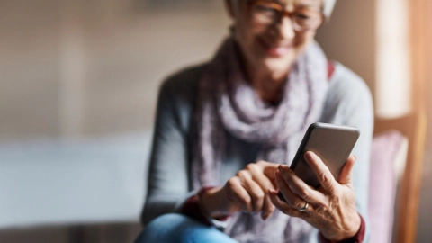 Sağlık uygulamasına sahip akıllı telefon (Kaynak: Getty Images/Cecilie_Arcurs) Gözlüklü yaşlı bir kadın kanepede oturuyor. Kadın sol elinde bir akıllı telefon tutuyor, sağ eliyle ekrana yazıyor ve gülümsüyor.