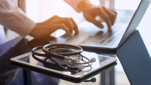 لابتوب وحاسب لوحي وسماعة طبية على طاولة زجاجية. شخص يكتب على لوحة مفاتيح اللابتوب.