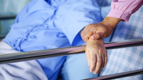 شخص مستلقٍ على سرير به درابزين معدني. ويضع ذراعه على درابزين السرير. وهناك شخص آخر يلمس ذراعه.