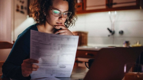 امرأة تجلس أمام لابتوب وتحمل في يدها ورقة معلومات.