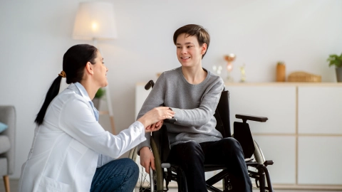 Bir doktor tekerlekli sandalyedeki bir çocuğun yanına diz çöküp ona elini uzatıyor.