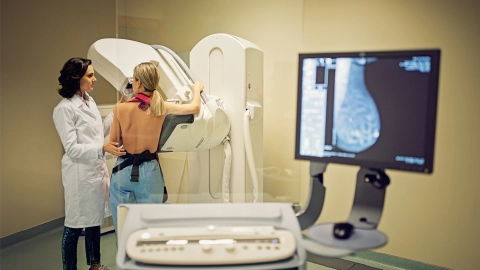 Cancer screening: patient undergoing mammography screening. A doctor is standing next to the woman. She is conducting the examination. The X-ray of a breast is visible on a screen.