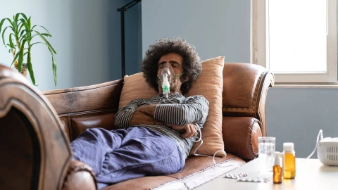 Мужчина лежит на диване. Он подключен к аппарату искусственной вентиляции легких через маску. На столе рядом с ним стоят лекарства.