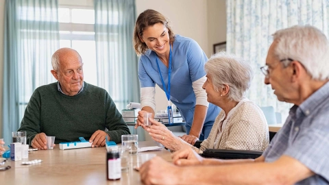 Older people seated at a table are being given medication by a nurse.