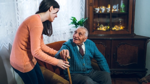 Yaşlı bir adam oturma odasındaki koltuğundan kalkmak istiyor, elinde bastonu var. Genç bir kadın ayağa kalkmasına yardım ediyor ve destek olarak kolunun altından tutuyor.