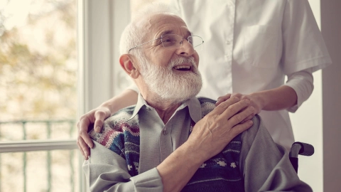 Care assessment: older man with a beard and glasses sitting in a wheelchair turning round smiling to a person in white standing behind him with his hands on his shoulders.