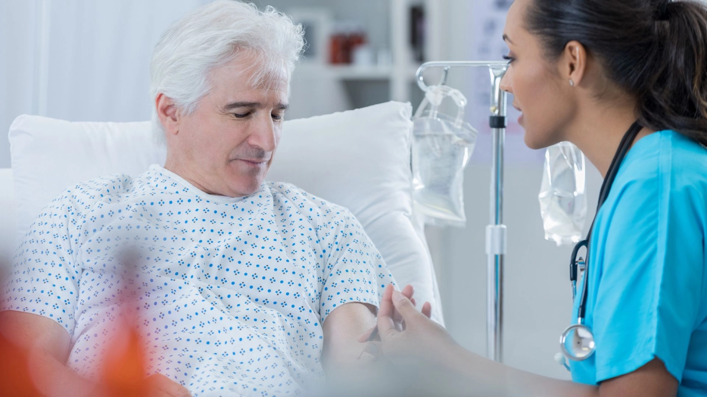 Patient und Krankenschwester (Symbolbild) (Quelle: Getty Images/SDI Productions) Prostatakrebs: Ein älterer Mann sitzt in einem Krankenhausbett. Er schaut nach unten, ihm gegenüber sitzt eine Krankenschwester. Sie schaut ihn an und entnimmt ihm Blut.