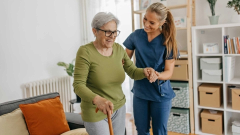 Bir hemşire yaşlı bir kadına destek oluyor. Yaşlı kadın baston kullanıyor. İkisi de gülümsüyor.