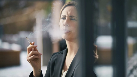 امرأة تقف بجانب النافذة وتمسك سيجارة وتخرج دخانًا من فمها.
