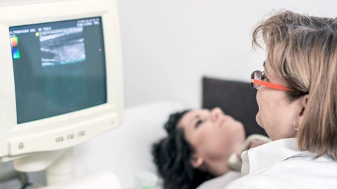 Thyroid cancer: patient lying on a hospital trolley bed (gurney). A doctor is sitting in front of her and taking an ultrasound scan of her thyroid gland. The doctor is examining the ultrasound image on a monitor.