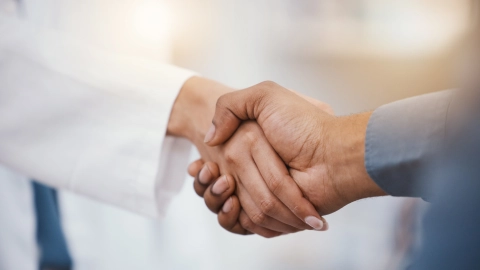 Doktor önlüğü giyen bir kişi ile gömlek giyen bir kişi arasında yapılan resmi el sıkışması.