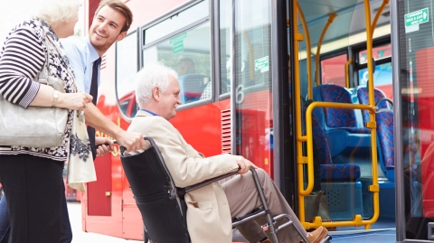 Genç bir otobüs şoförü tekerlekli sandalyedeki yaşlı bir adamı rampadan iterek otobüse bindiriyor.