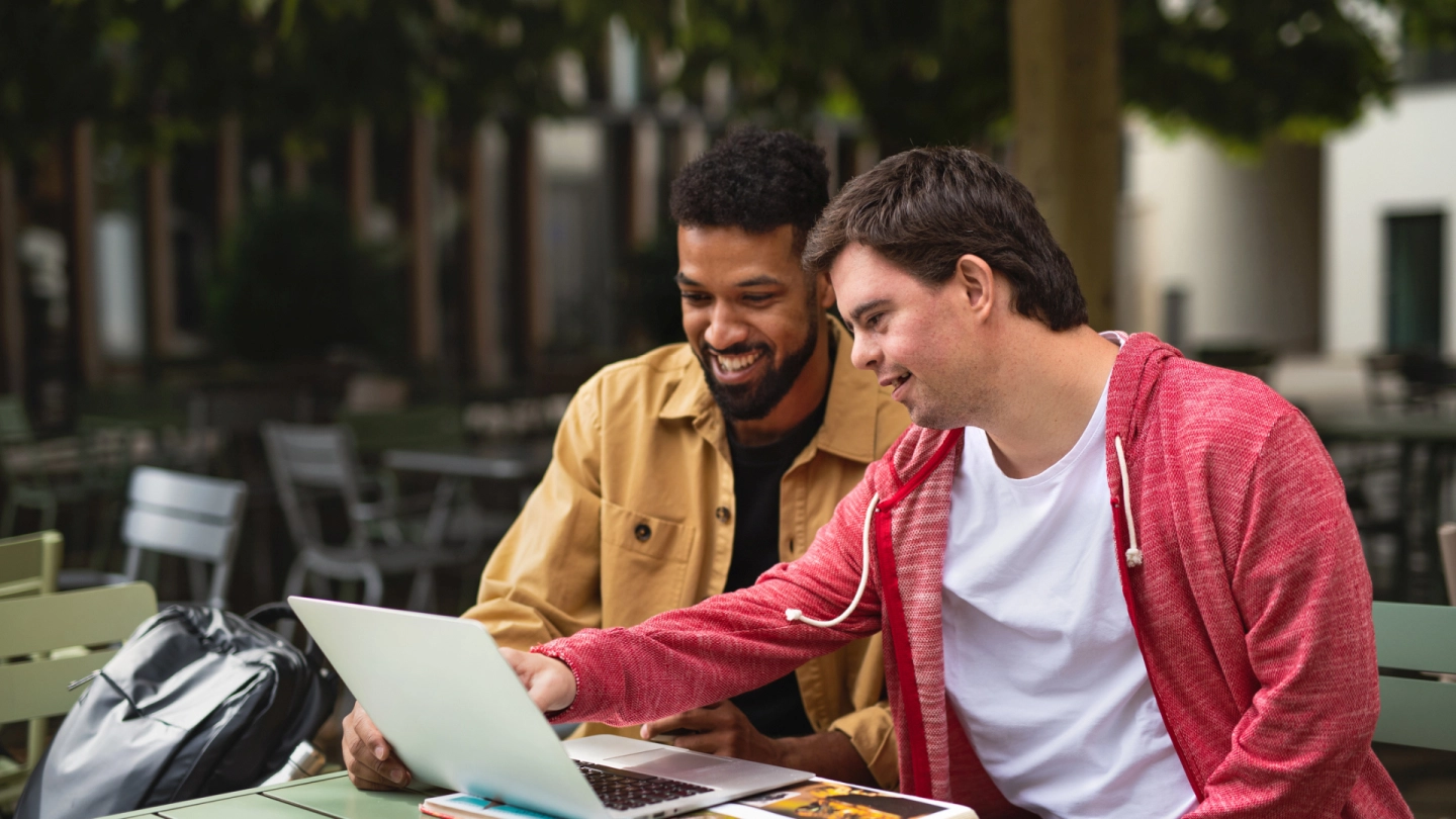 Leistungen zur Teilhabe (Quelle: iStock/Halfpoint) Ein junger Mann mit Behinderung und ein weiterer junger Mann schauen gemeinsam auf einen Laptop.