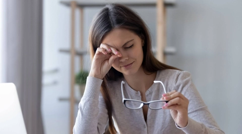 Женщина сняла очки и потирает глаза. Рядом на столе стоит ноутбук.