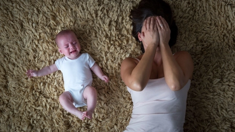 Depressive Mutter mit Kind (Quelle: Getty Images/SolStock) Eine Frau liegt auf einem Teppich und hält sich beide Hände vors Gesicht. Sie wirkt erschöpft. Neben ihr liegt ein Baby. Es scheint zu strampeln und zu weinen.